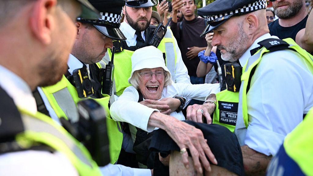 Police officers surround and arrest an old woman who appears to be in distress as they lift her off the ground. Police officers surround and arrest an old woman who appears to be in distress as they lift her off the ground.
